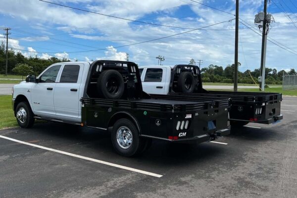 A white crew cab truck with a black flatbed spare tire and tool compartments viewed from left rear